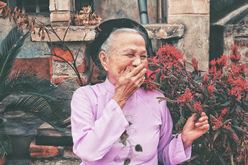 An elderly woman sitting next to red flowers and smiling.